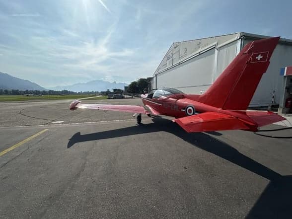 A red aircraft, HB-EML, parked on the tarmac with other aircraft in the background.