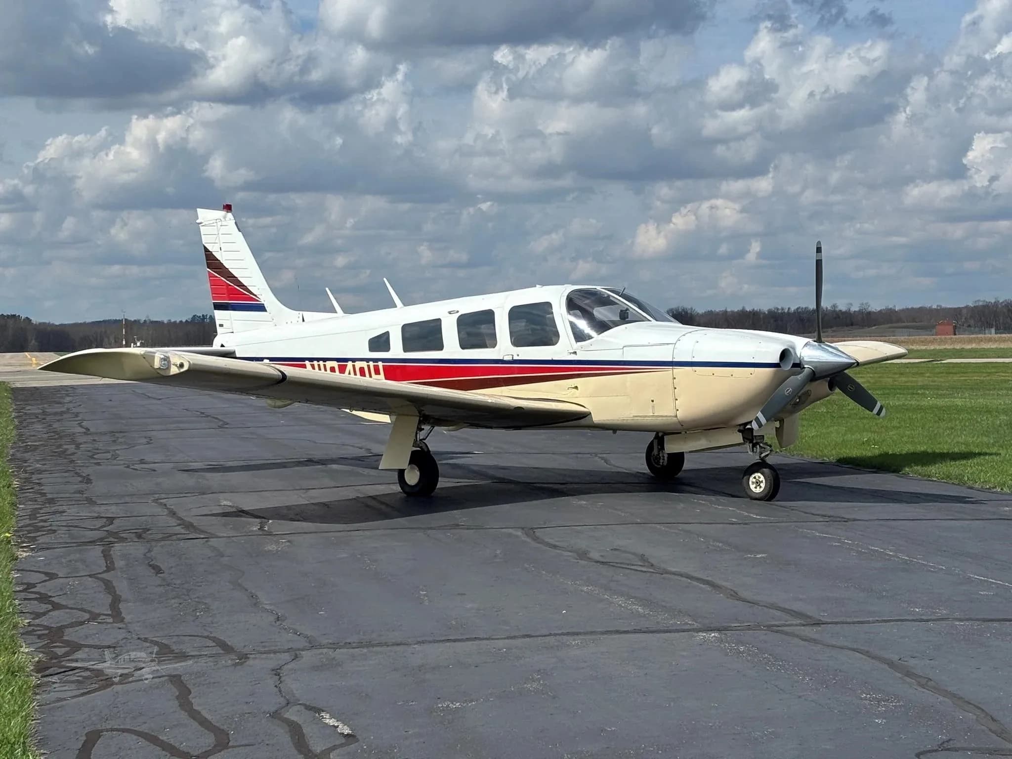 A white and beige single-engine aircraft with red and blue stripes on the tail and fuselage, parked on an asphalt runway under a cloudy sky.