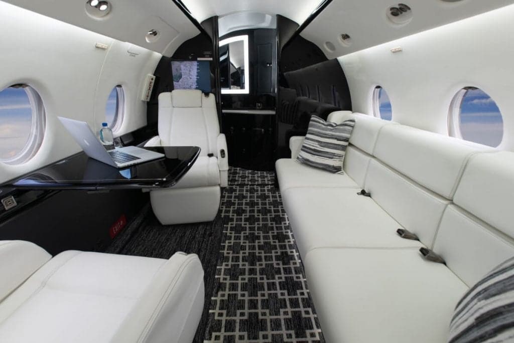 View of the aircraft's interior with white leather seats, a sofa, and a table with a laptop.