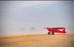 A red Cubcrafters aircraft is parked on a dirt runway in foggy conditions.