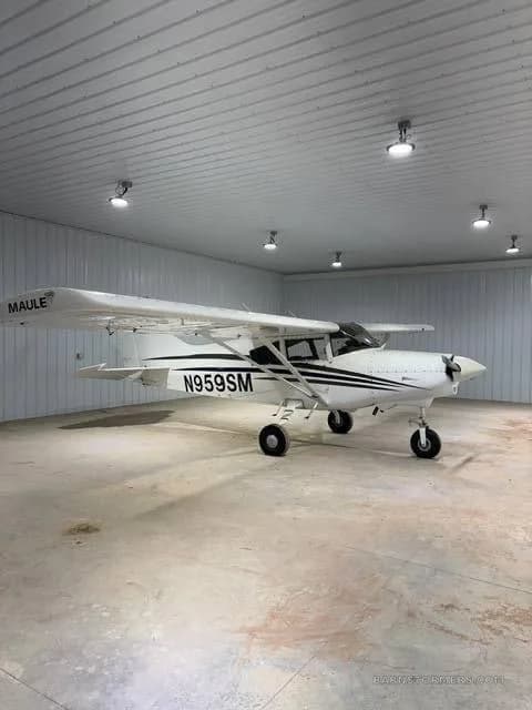 A white Maule M-7-235B aircraft with black stripes and registration N959SM is parked inside a hangar.