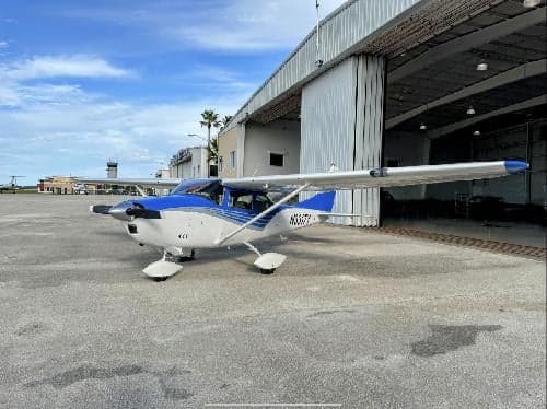 A Cessna 182 Skylane aircraft with a blue and white paint scheme is parked on the tarmac in front of a hangar.