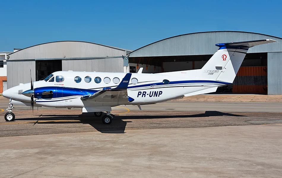 A white Beechcraft King Air 350 with blue and dark blue stripes is parked on a tarmac next to a hangar under a clear blue sky.