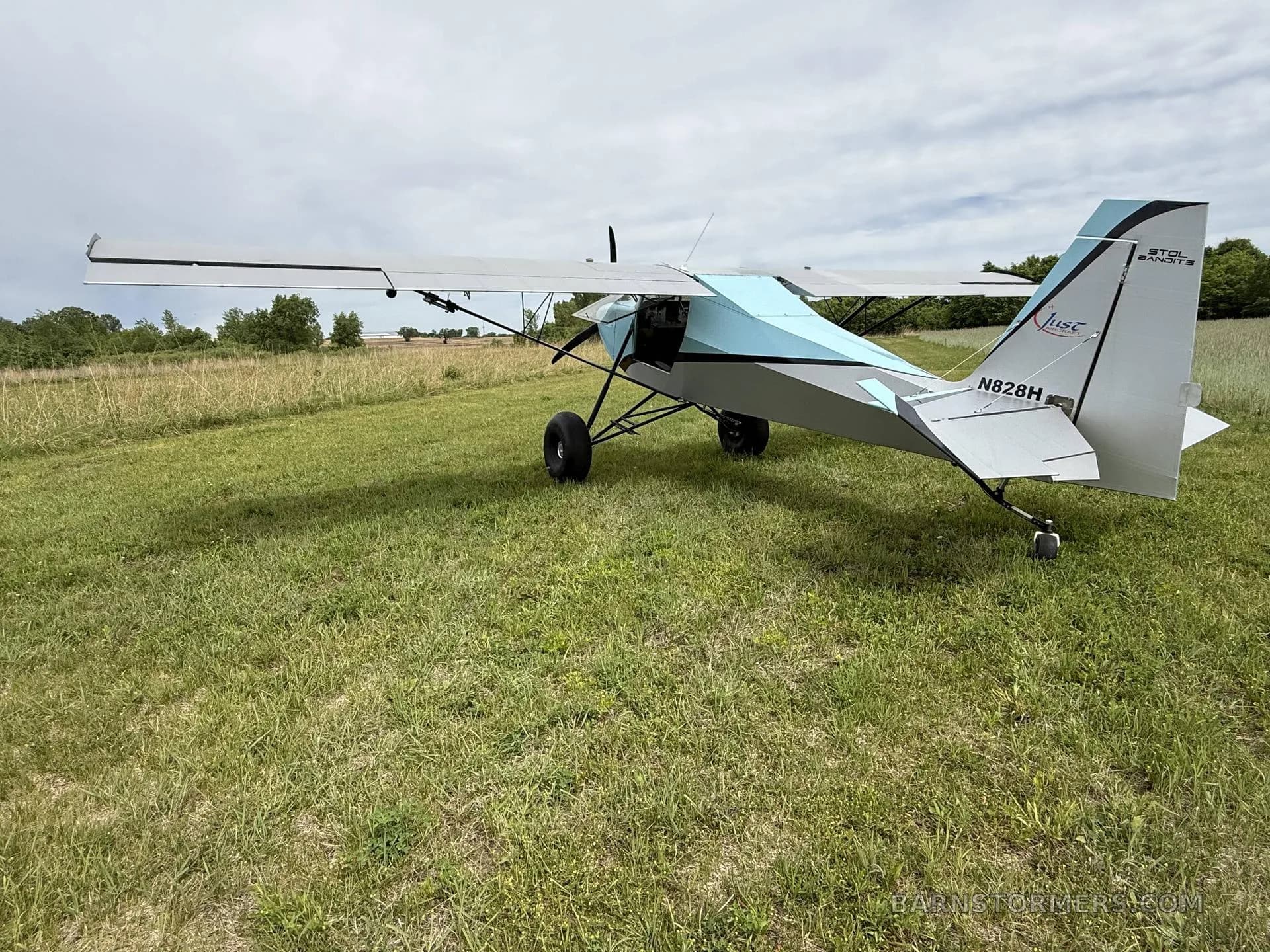 A rear-left view of a light blue and grey Just Aircraft SuperSTOL with its wings folded, parked on a grassy field.
