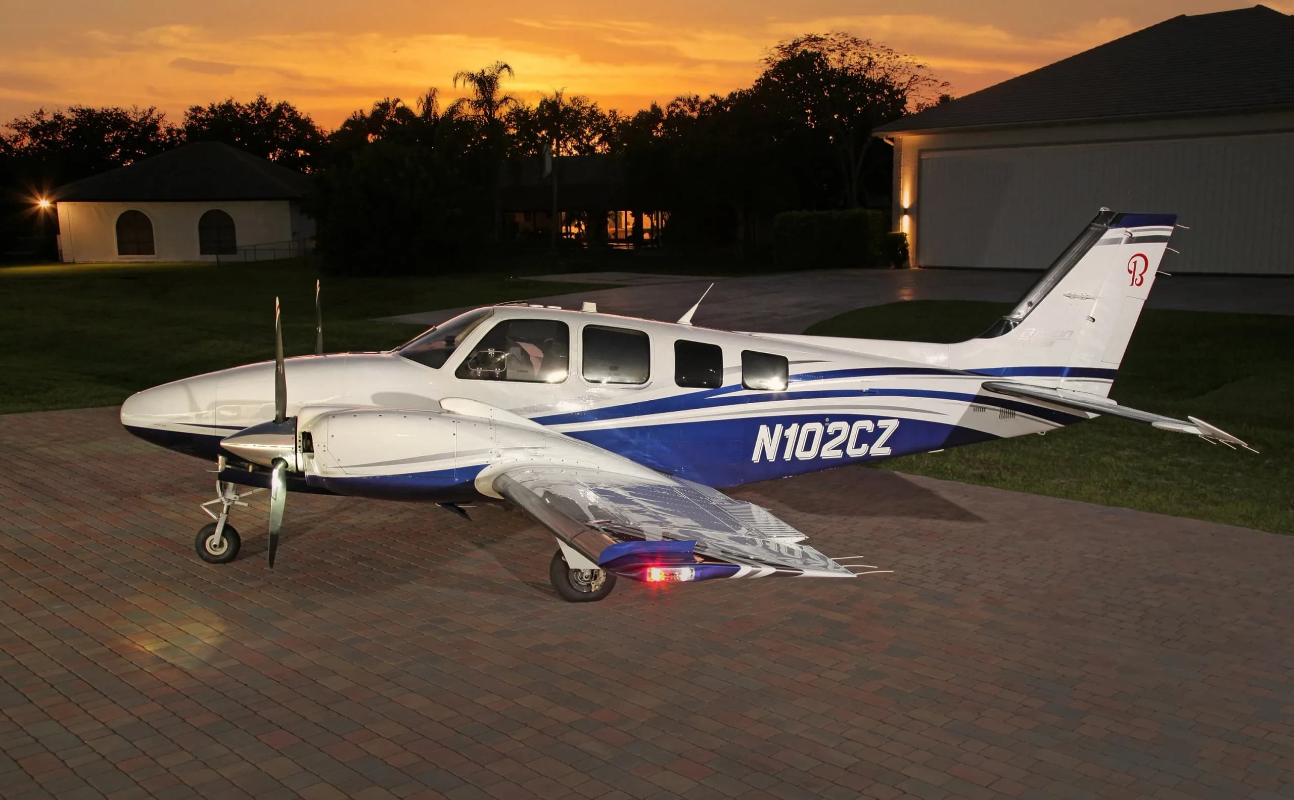 A white and blue Beechcraft Baron N102CZ parked on a paved driveway at sunset.