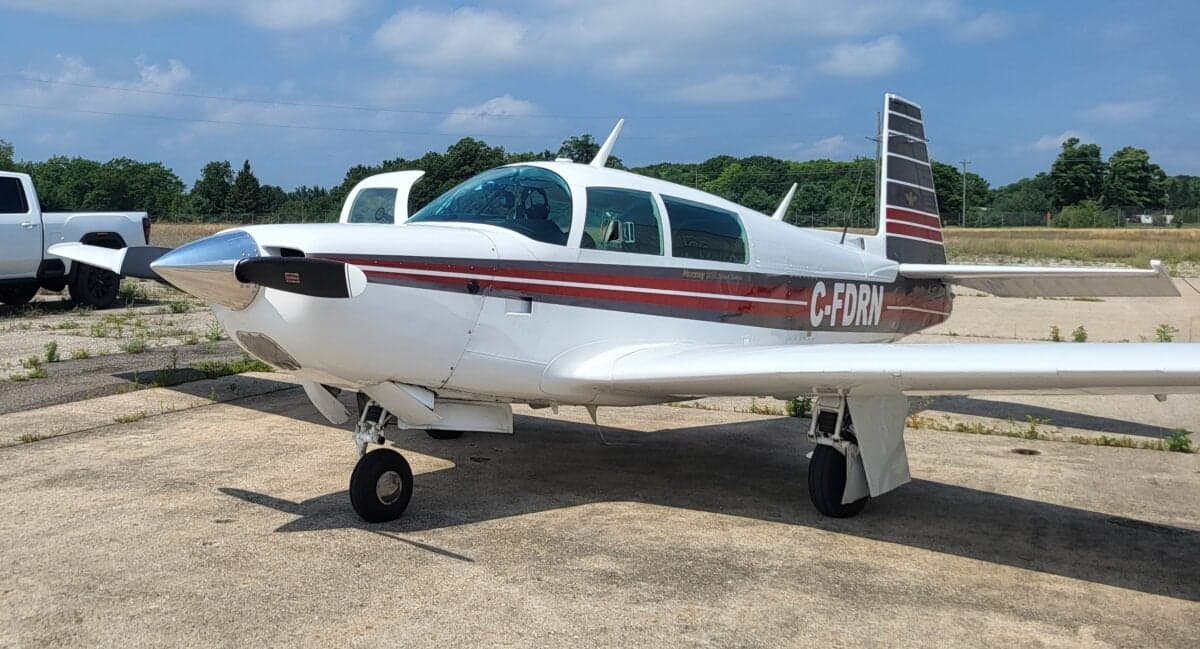 A white Mooney M20J aircraft with red and grey stripes, registration C-FDRN, parked on an asphalt ramp.