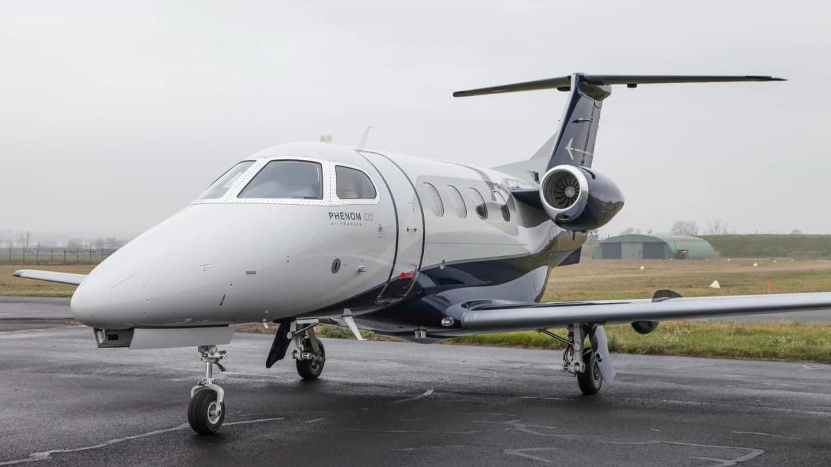 A white Embraer Phenom 100 with dark blue and grey livery parked on a wet tarmac.