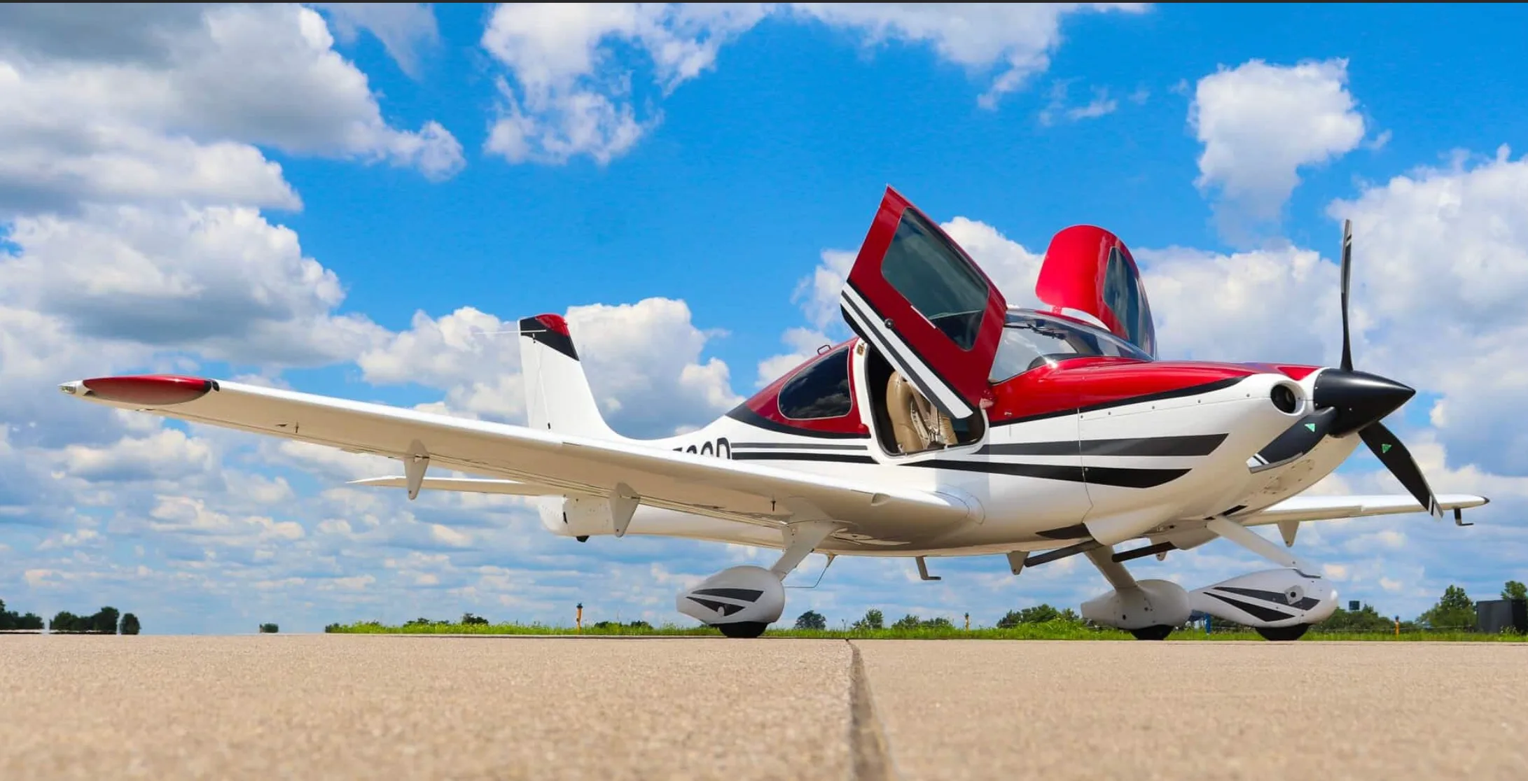 A white and red single-engine aircraft with open gull-wing doors sits on an airfield under a blue sky with clouds.