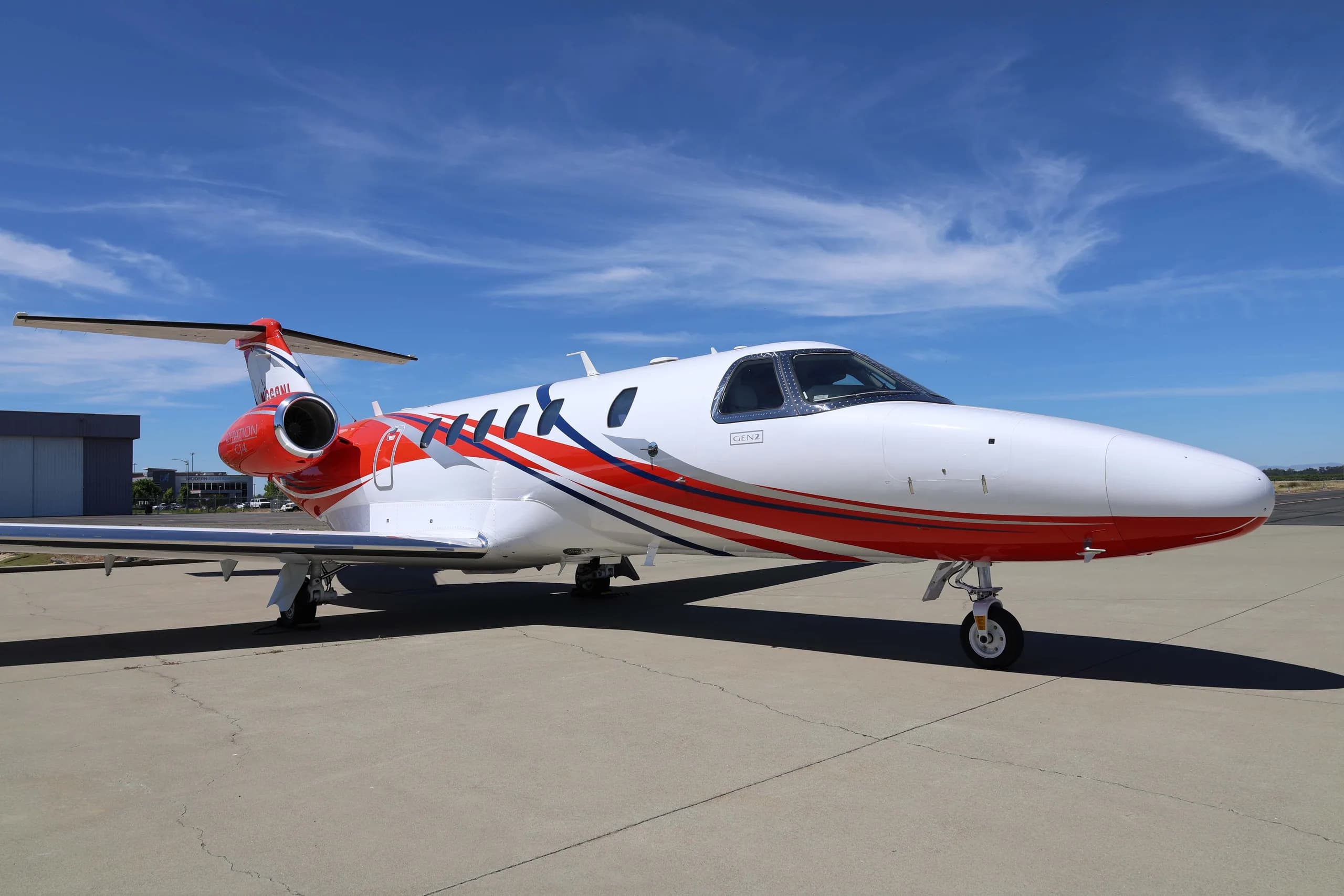 A white Cessna Citation CJ4 (N369NL) with red and blue stripes on the fuselage, parked on an airfield under a blue sky.
