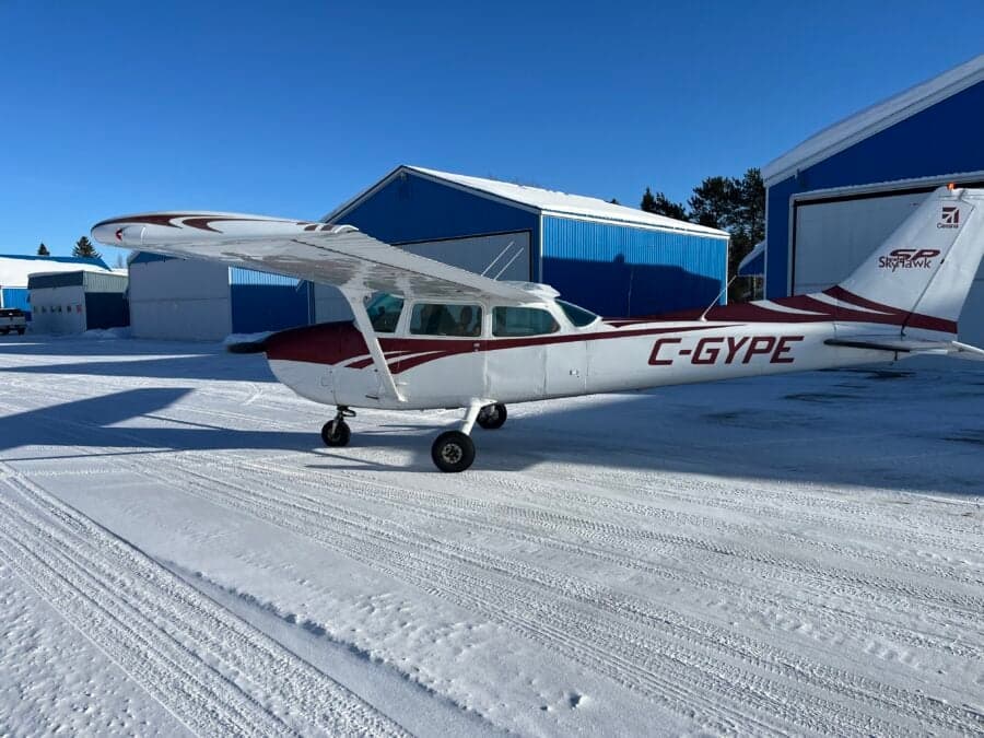 A white and red Cessna 172 (C-GYPE) parked in a snowy field with blue hangars in the background.