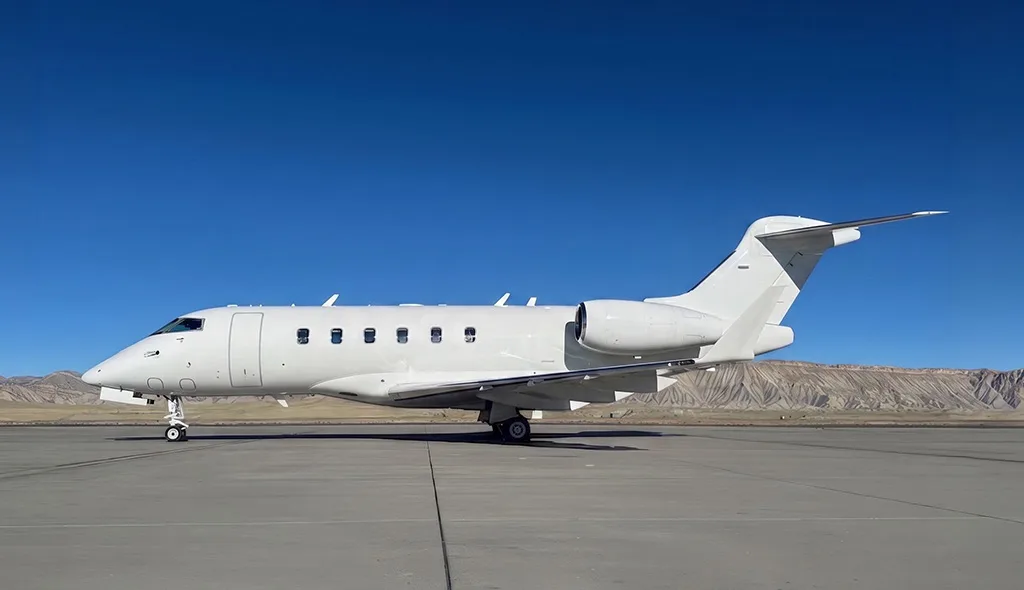 A white Bombardier Challenger 605 business jet parked on an airfield with mountains in the background.