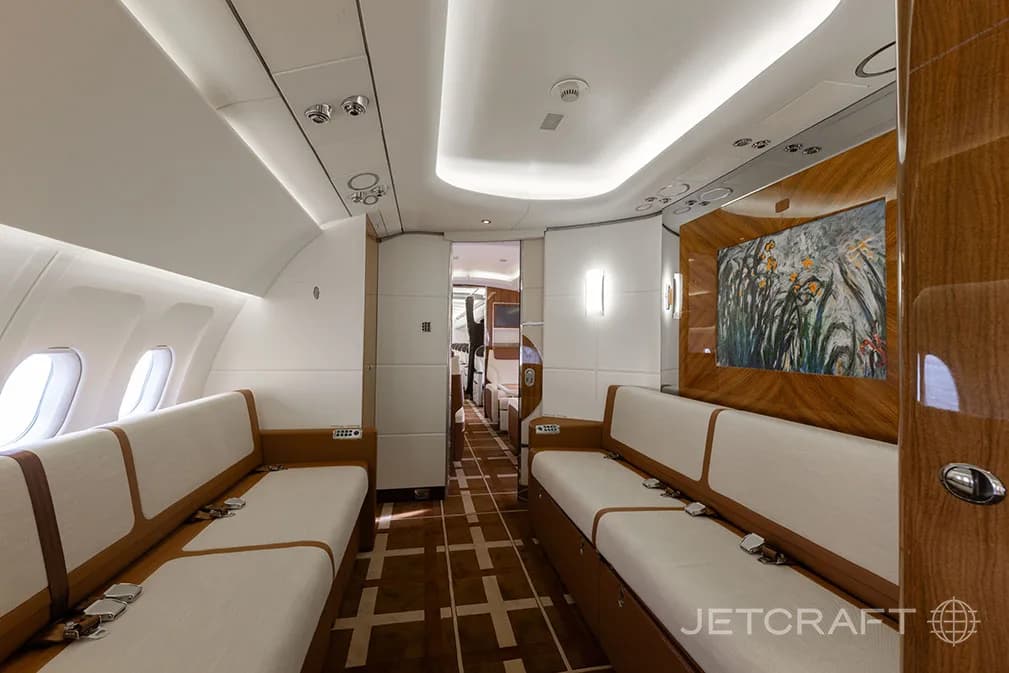A luxurious aircraft cabin interior features two long white couches facing each other, with a wooden wall panel displaying artwork.