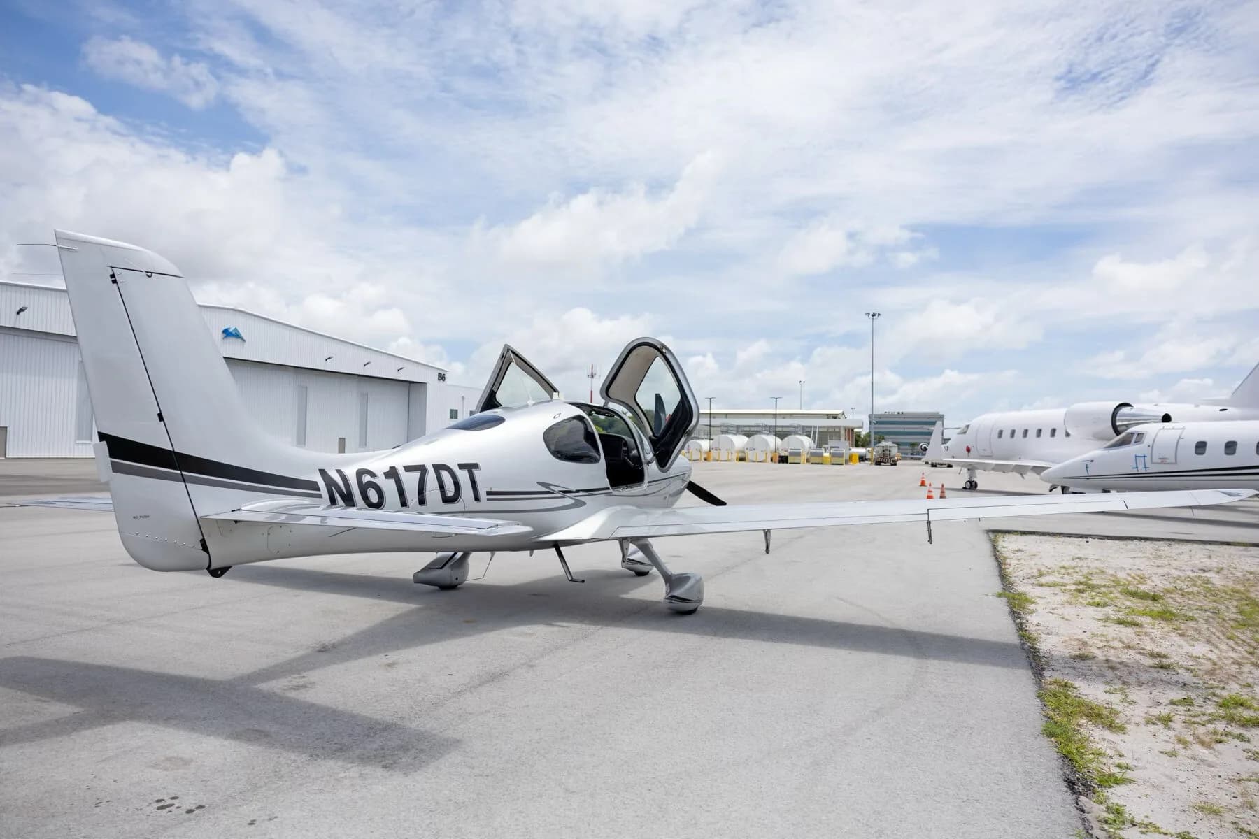 A silver Cirrus SR22 aircraft with its gull-wing doors open, parked on an tarmac next to other aircraft.