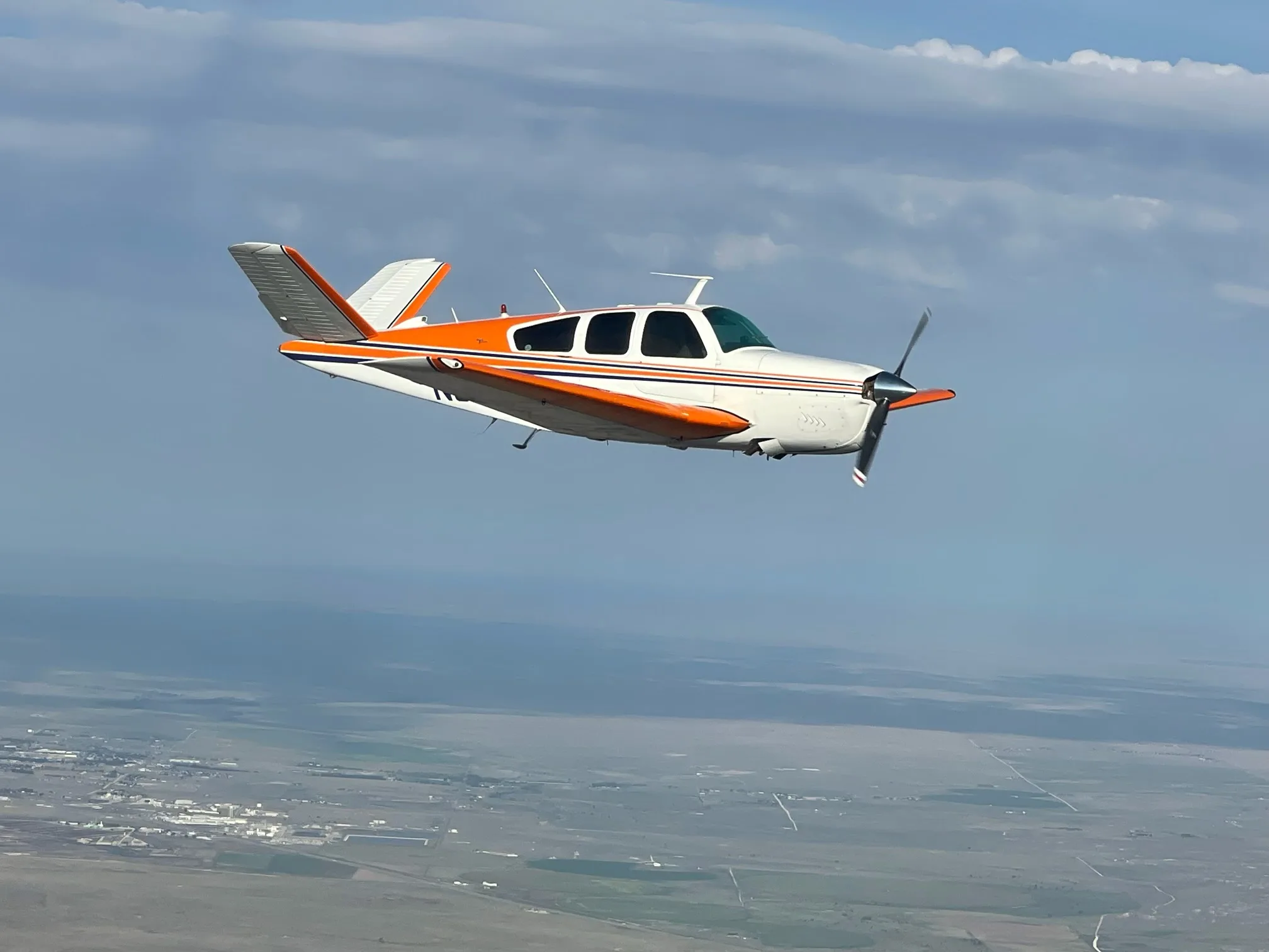 A Beechcraft V35 Bonanza aircraft in flight with an orange and white paint scheme.