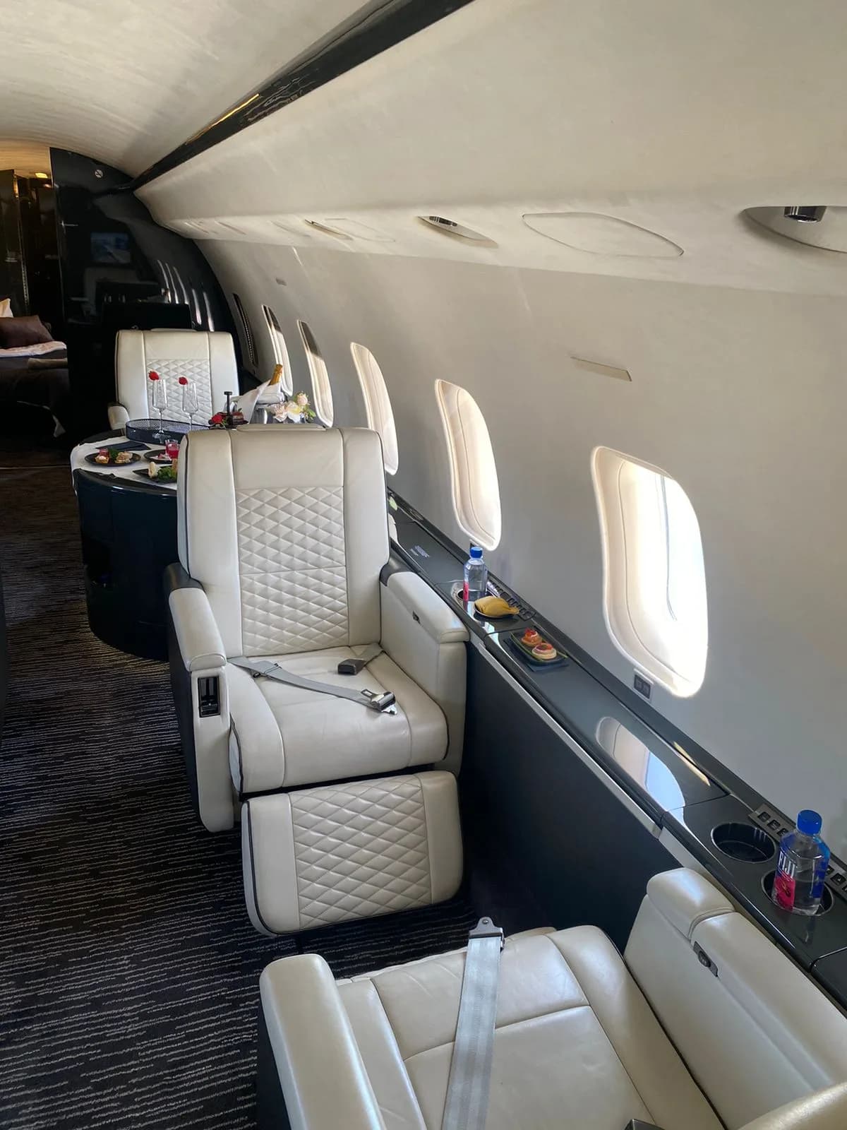 A luxurious private jet interior with white quilted leather seats and a table set with food and drinks.