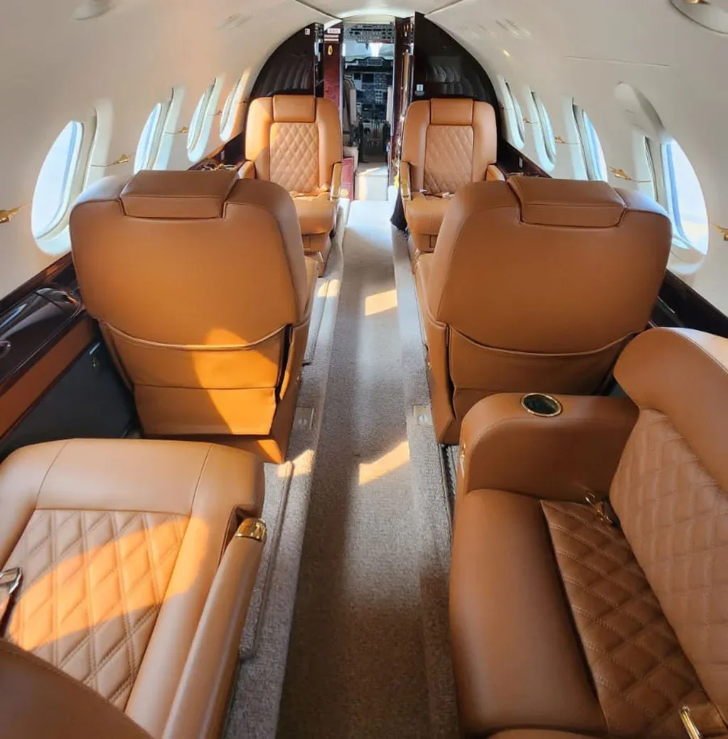 A long view down the aisle of a business jet cabin, showing several brown leather seats and the cockpit entrance in the distance.