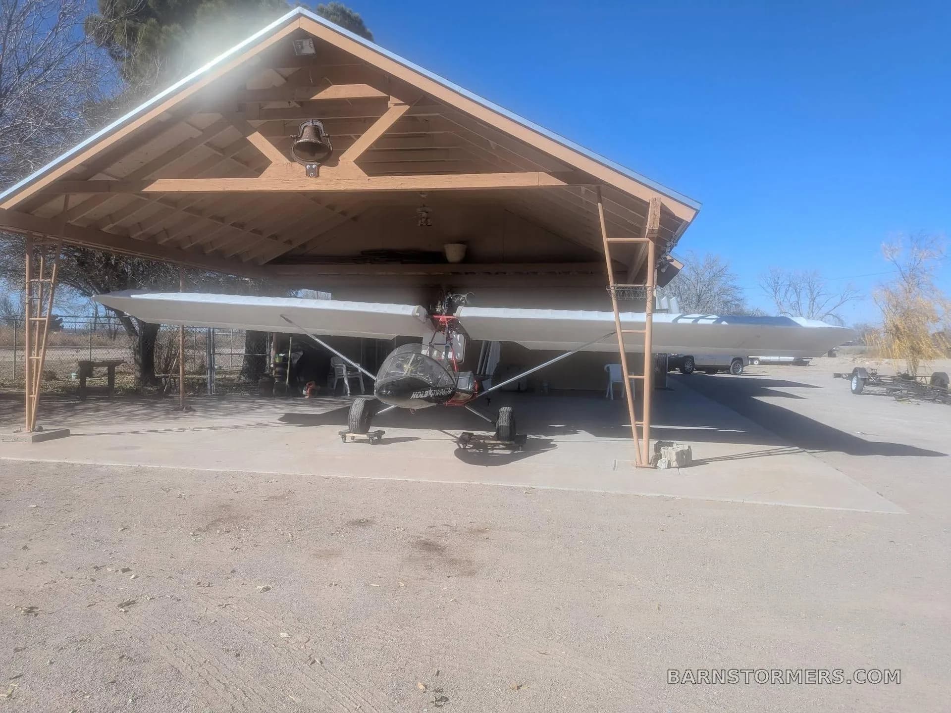 A black and white Volmer VJ-24W SunFun experimental aircraft is parked under a wooden carport on a sunny day.