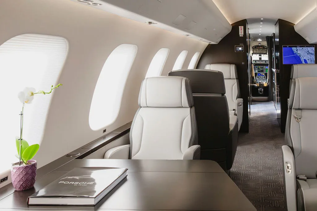 A view inside a private jet's cabin, showing light grey seats, a table with a book, and the cockpit visible in the distance.