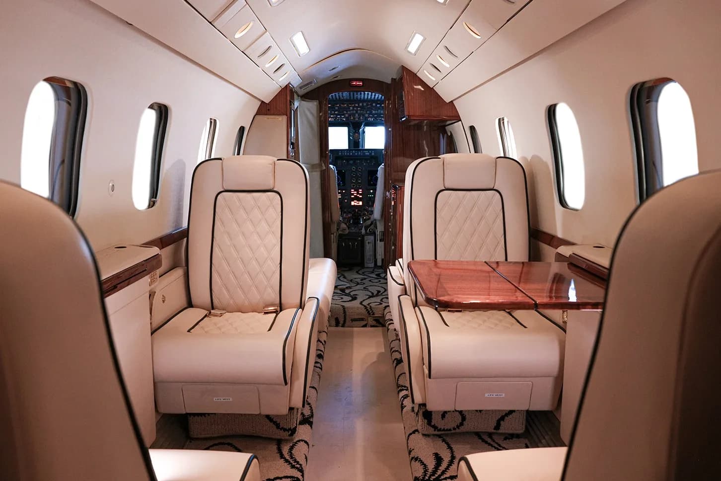 A view down the aisle of a private jet cabin, showing cream leather seats, a wooden table, and the cockpit beyond.