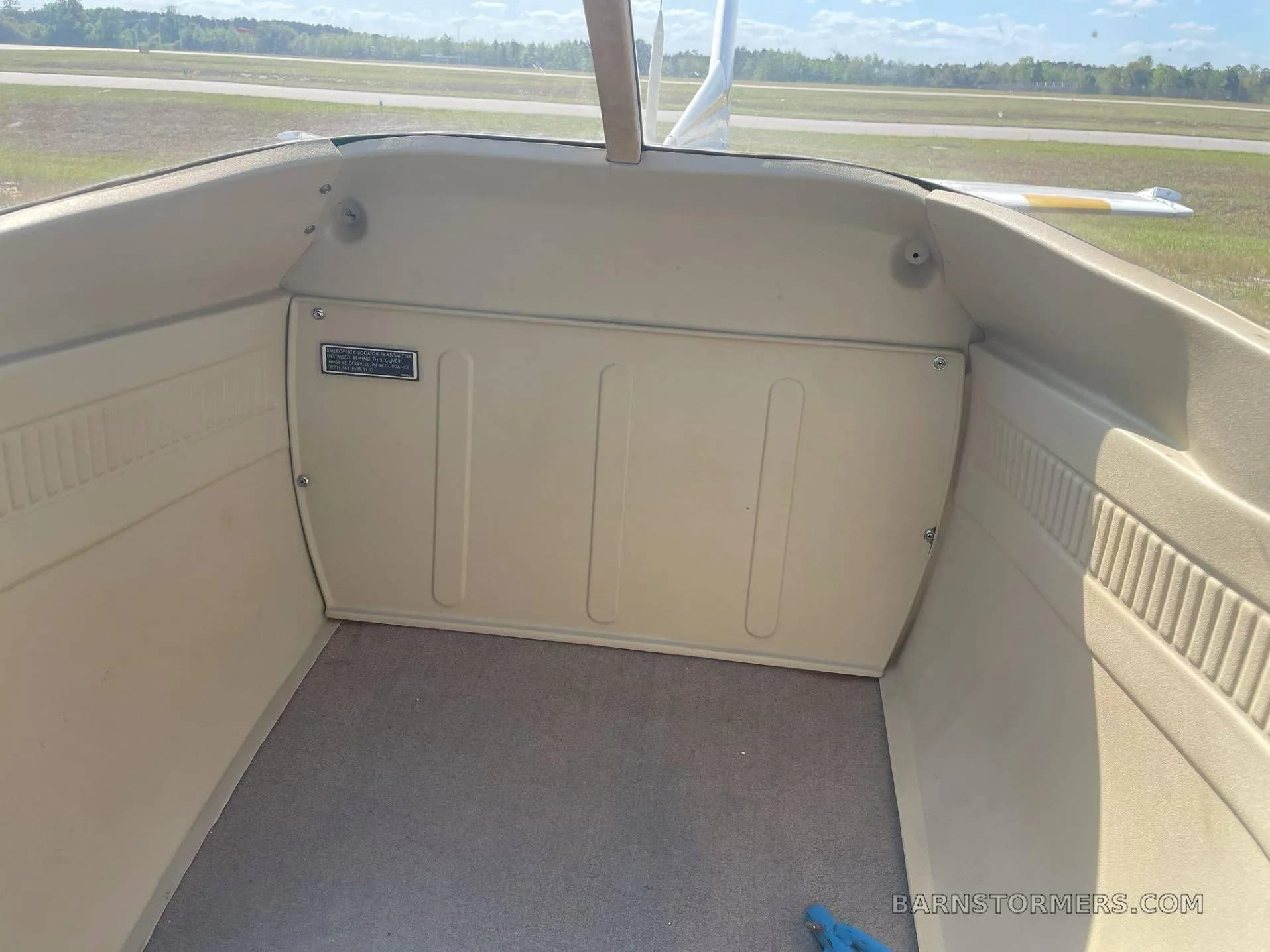 The beige-colored baggage compartment of a small aircraft is visible, with an airfield in the background through the rear window.