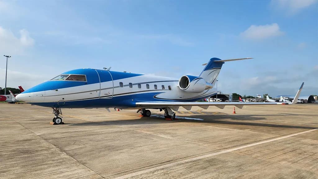 A blue and white Bombardier Challenger 604 business jet is parked on an airport tarmac under a clear blue sky.
