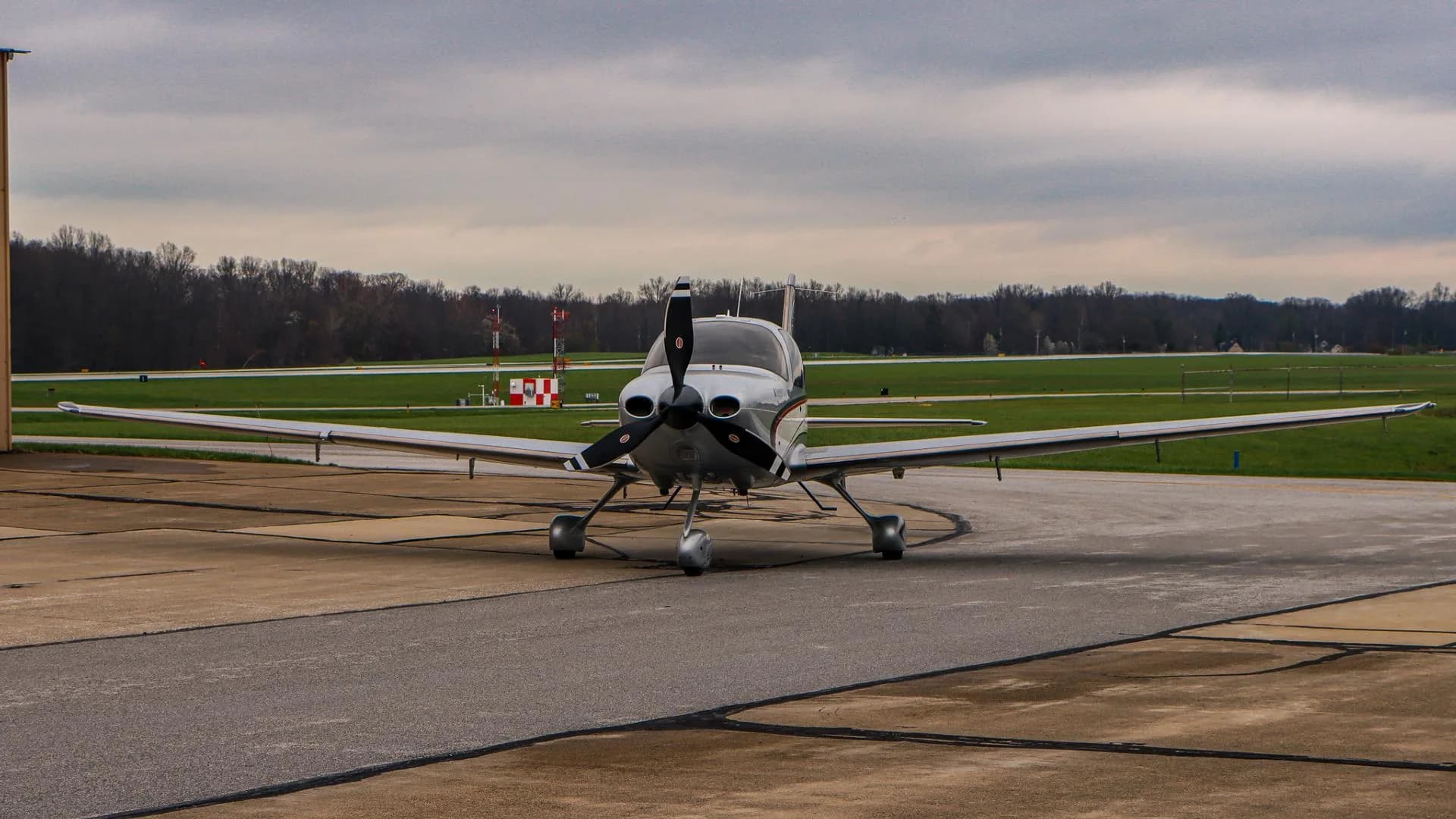 A silver and white Cirrus SR22 aircraft is seen from the front, parked on an airport tarmac with distant airport structures under a cloudy sky.