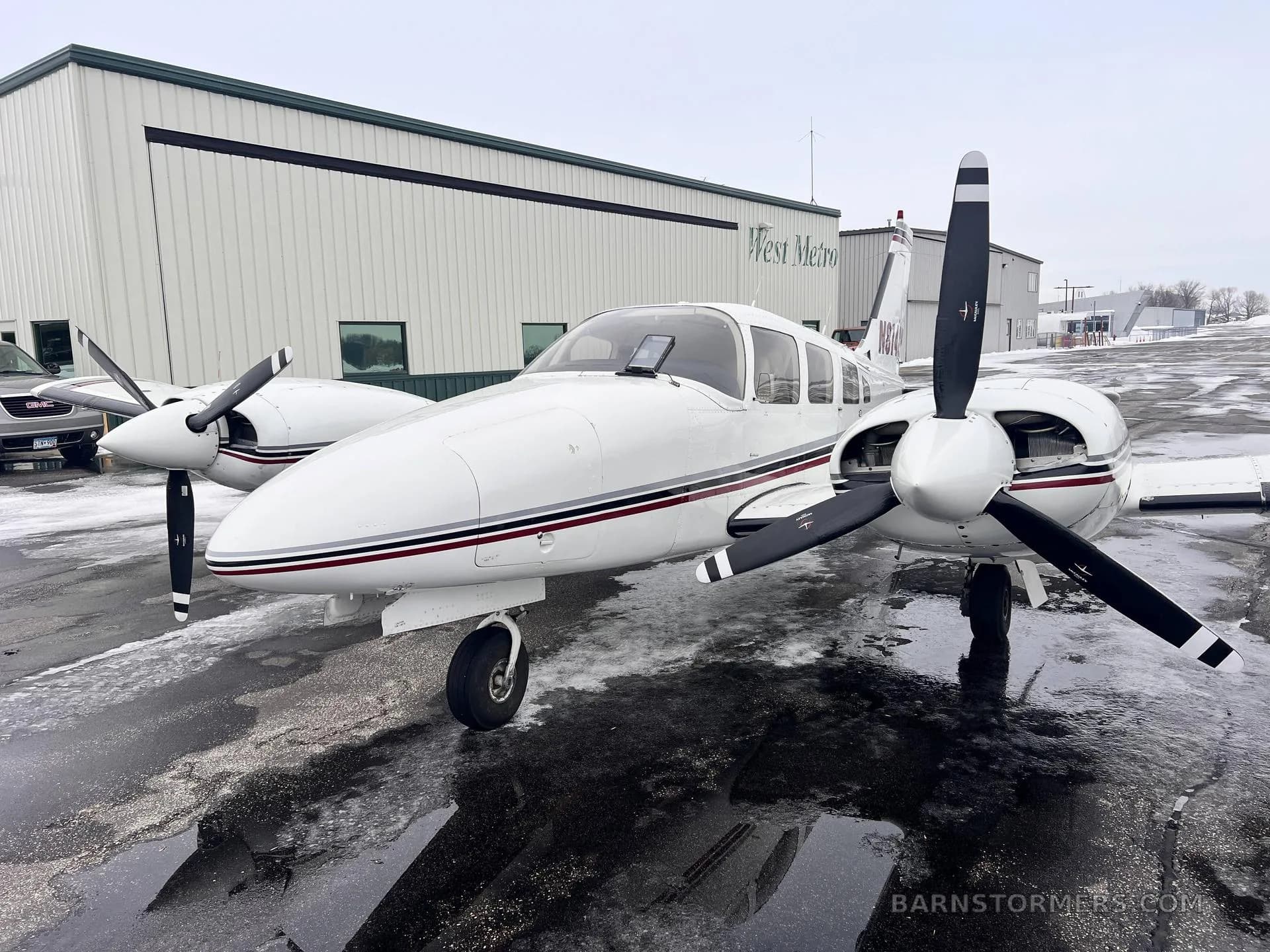 A front-quarter view of a white twin-engine Piper aircraft with maroon and black stripes, tail number N814CH, parked on a wet tarmac in front of a hangar.