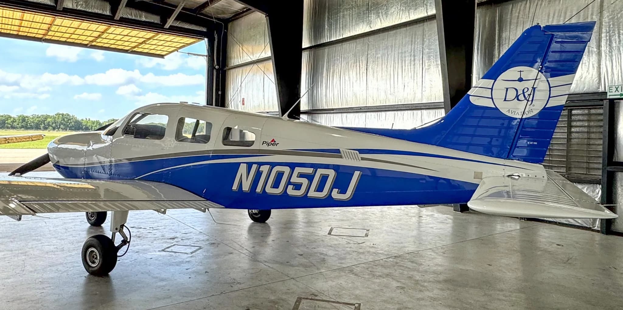 The white and blue Piper Archer TX, N105DJ, is parked in a hangar, seen from the rear-left with an open hangar door.