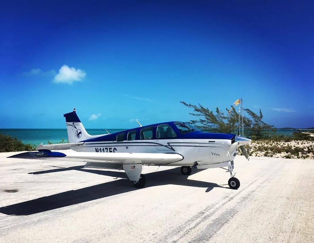 A white and blue single-engine aircraft, N117EC, is parked on a sandy strip next to a tropical beach under a clear blue sky.