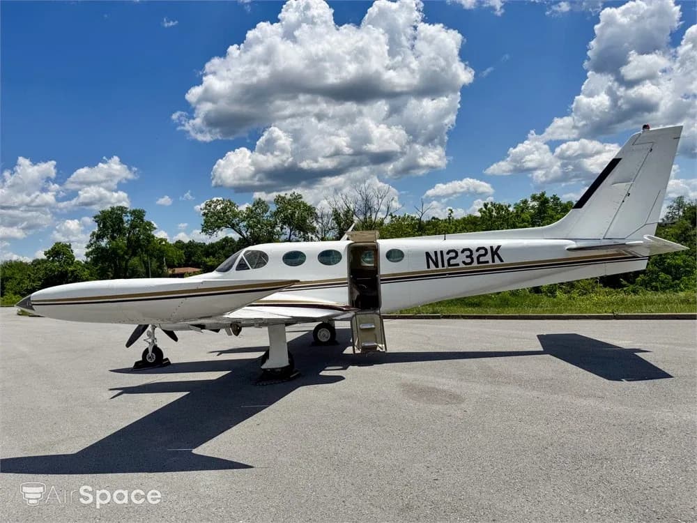 A white twin-engine aircraft with gold and brown stripes and its passenger door open, parked on an asphalt ramp.