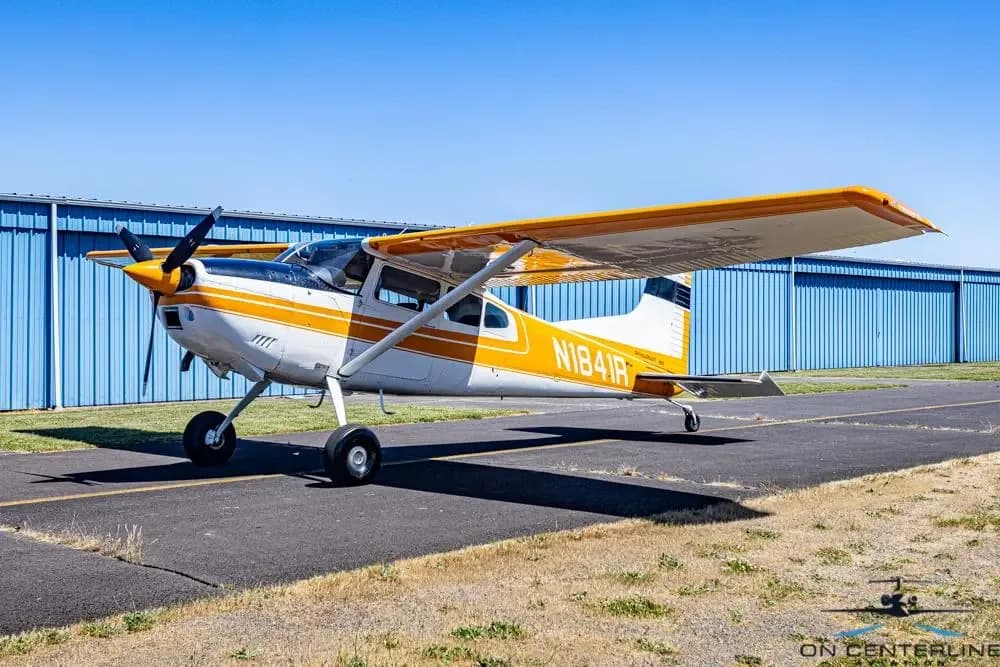 A white and orange Cessna 185 Skywagon (N1841R) parked on an asphalt ramp with blue hangars in the background.