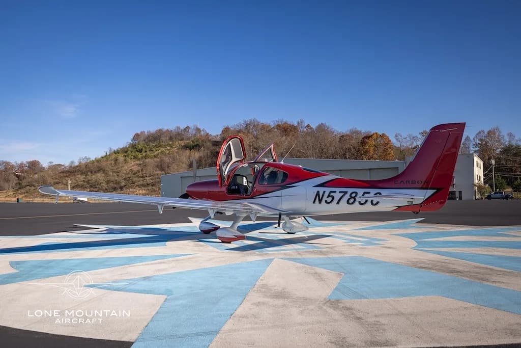A red and white Cirrus SR22 aircraft with registration N5785C parked on an airfield with its doors open, a hill in the background.