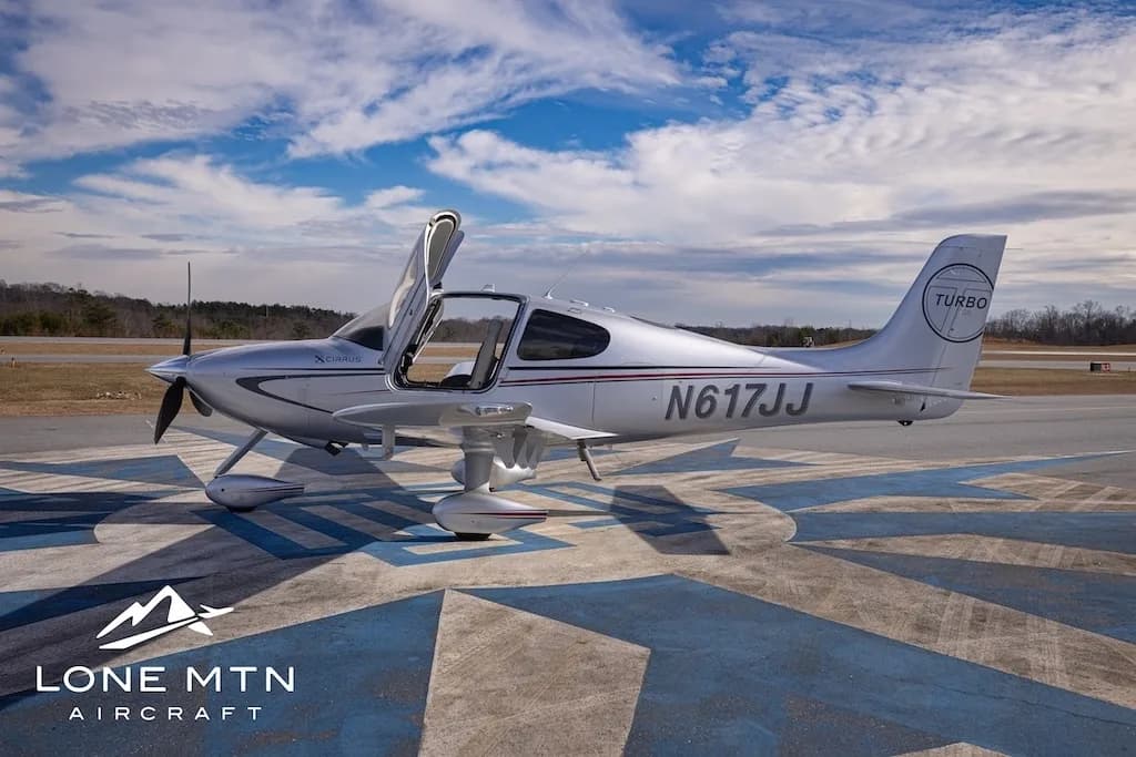 A silver Cirrus SR22T aircraft, N617JJ, with its doors open, parked on an airfield from a side view.