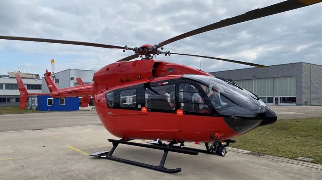 A red Airbus H135 helicopter with black skids and tinted windows parked on an airfield under a cloudy sky.