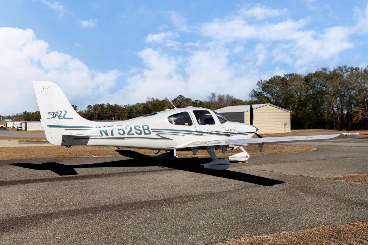 A side view of the white Cirrus SR22 aircraft, registration N752SB, parked on an asphalt ramp with a small building and trees in the background.