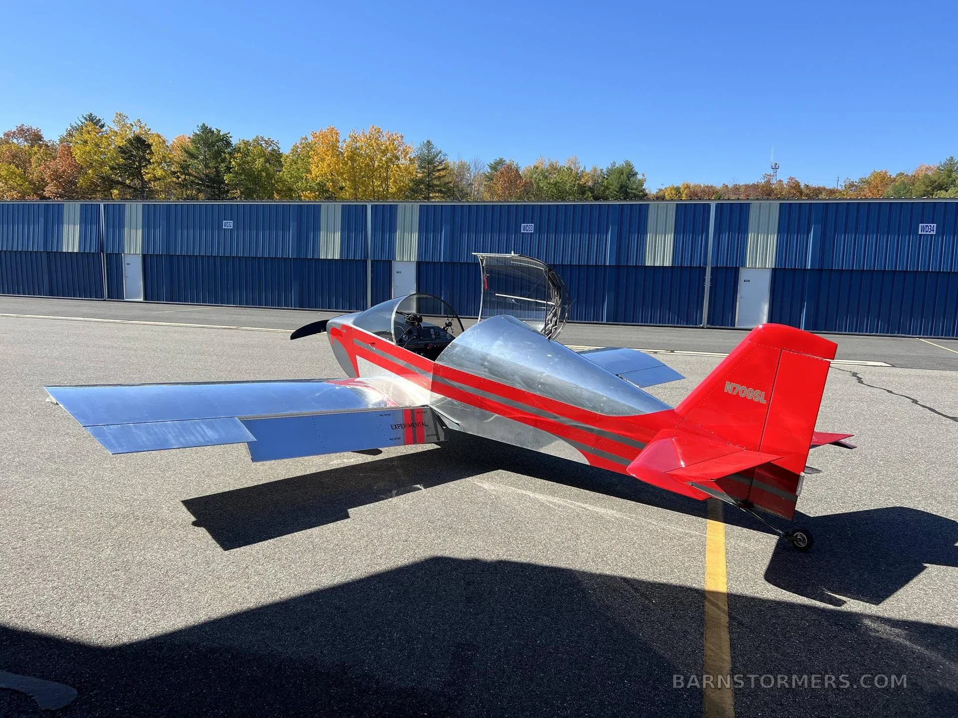 A red and silver experimental aircraft with an open canopy is parked on an asphalt apron in front of blue storage units.