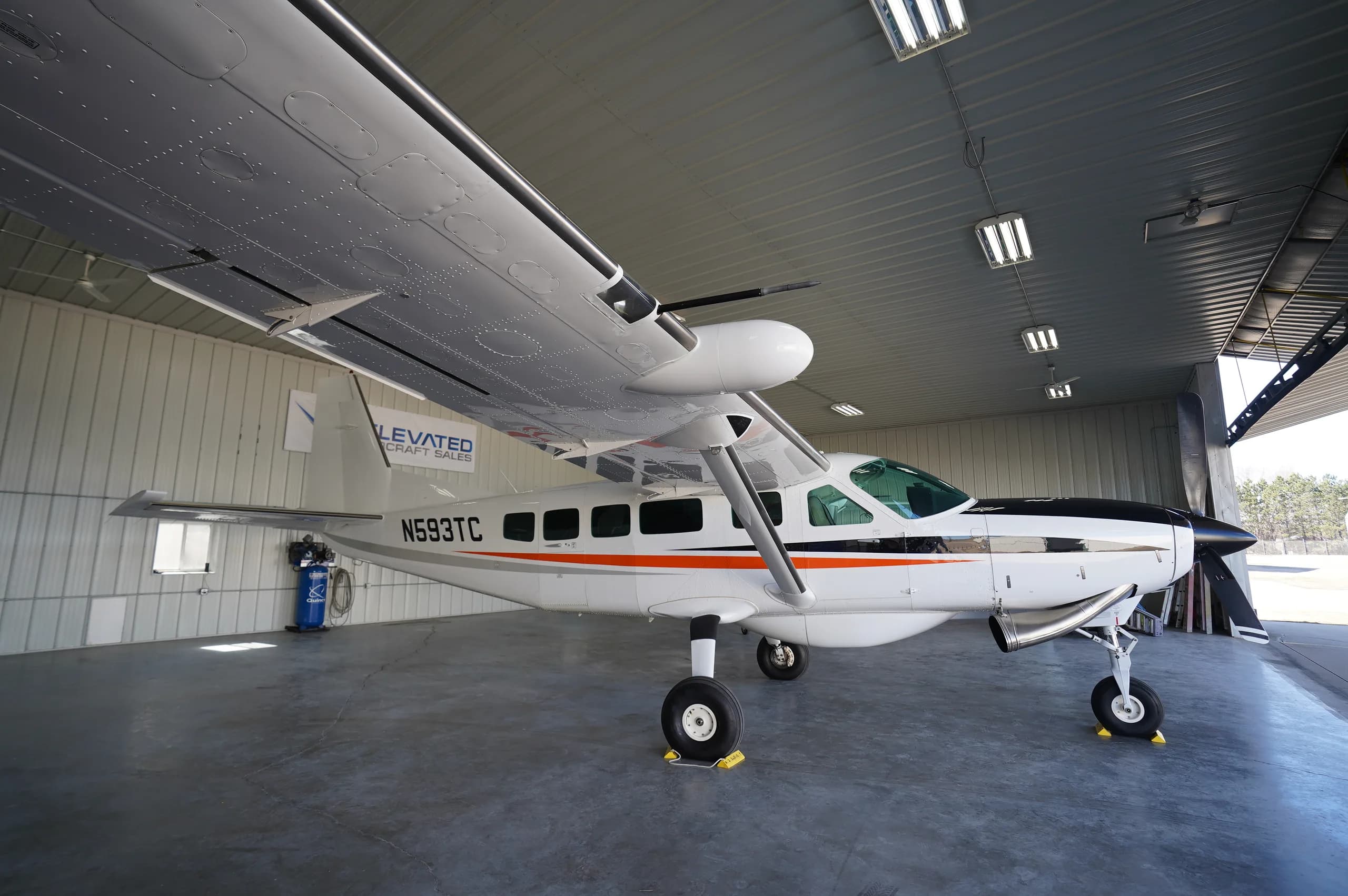 A white Cessna Caravan (N593TC) with black and orange stripes parked inside a hangar, viewed from the rear-right.
