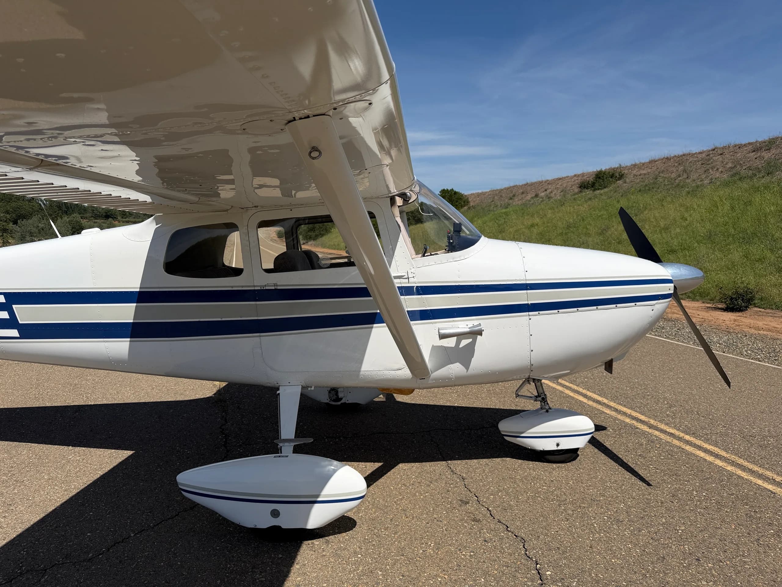 A close-up side view of the front fuselage of a white Cessna 172, showing the propeller, landing gear, and blue stripe against a blue sky.