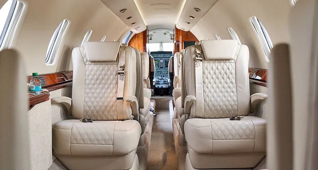 A view down the aisle of a private jet cabin, showing light-colored quilted leather seats and the cockpit visible in the distance.
