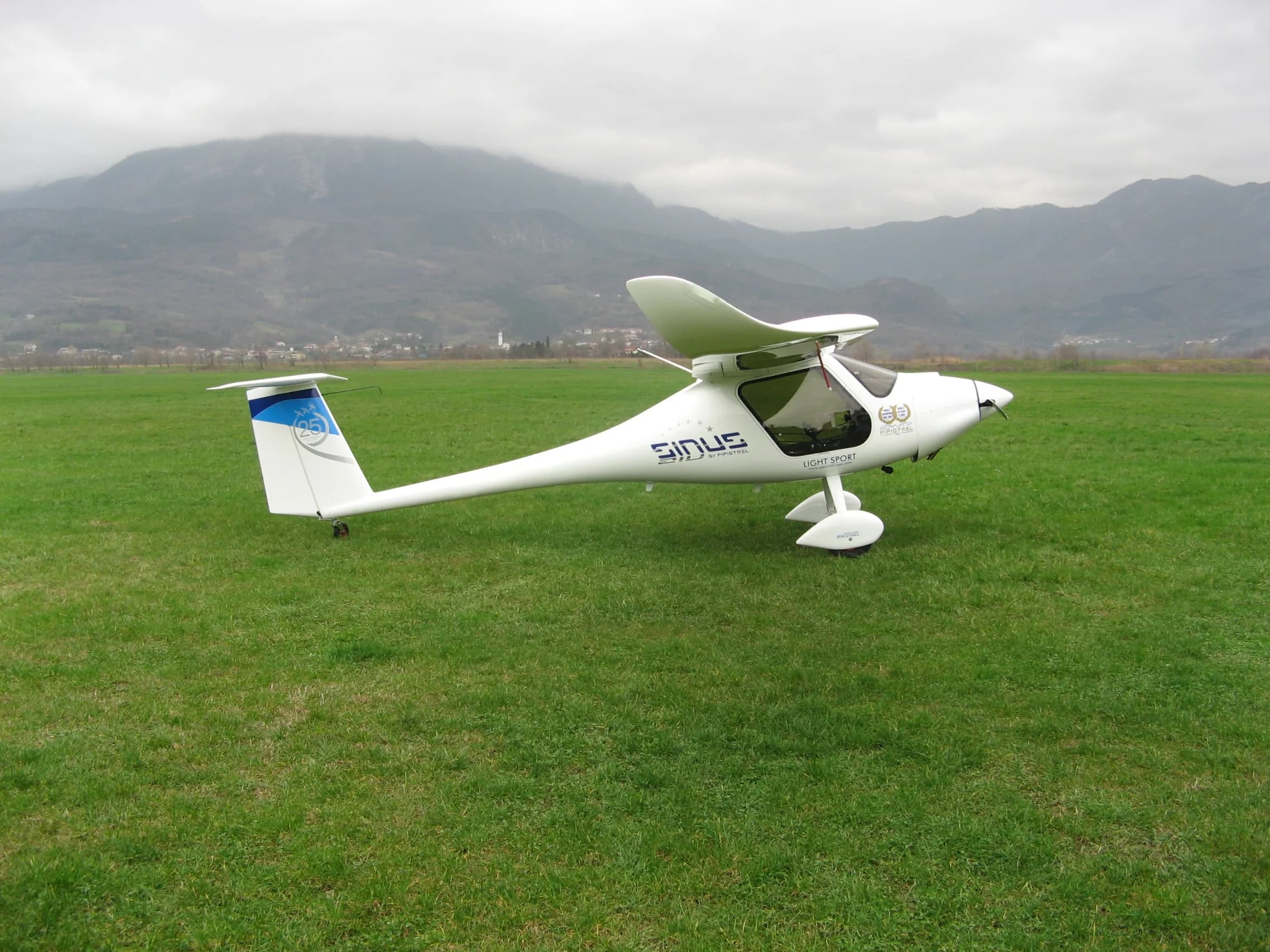 A white Pipistrel Sinus light sport aircraft with blue tail accents is parked on a grassy field, with mountains and a village in the background under a cloudy sky.