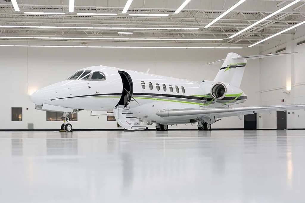 A white business jet with green and black stripes parked in a brightly lit hangar, with its main door open and stairs extended.