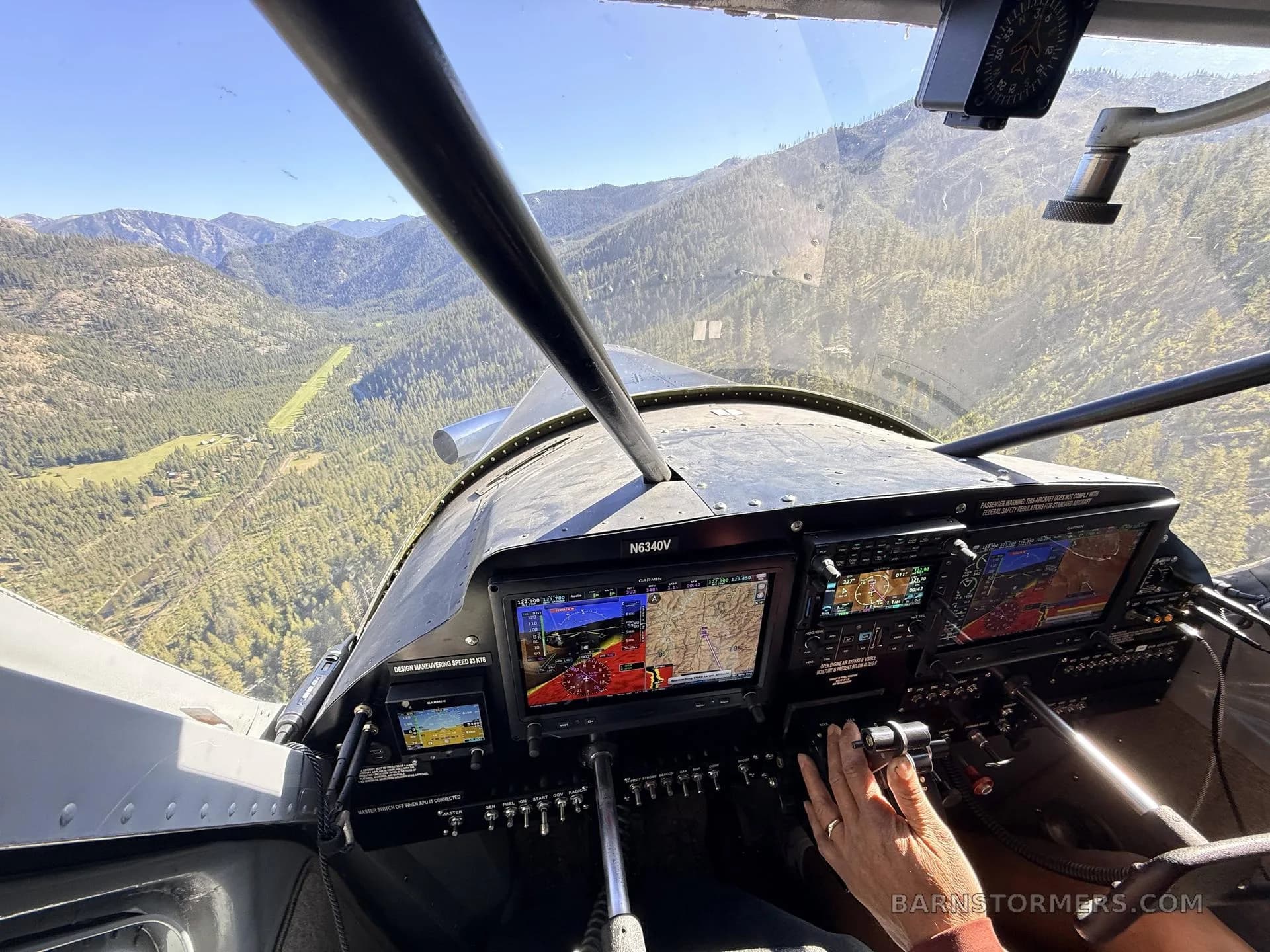 The cockpit of the T-MCX aircraft (N634DV) shows multiple digital displays and controls, with a view of forested mountains through the windshield.