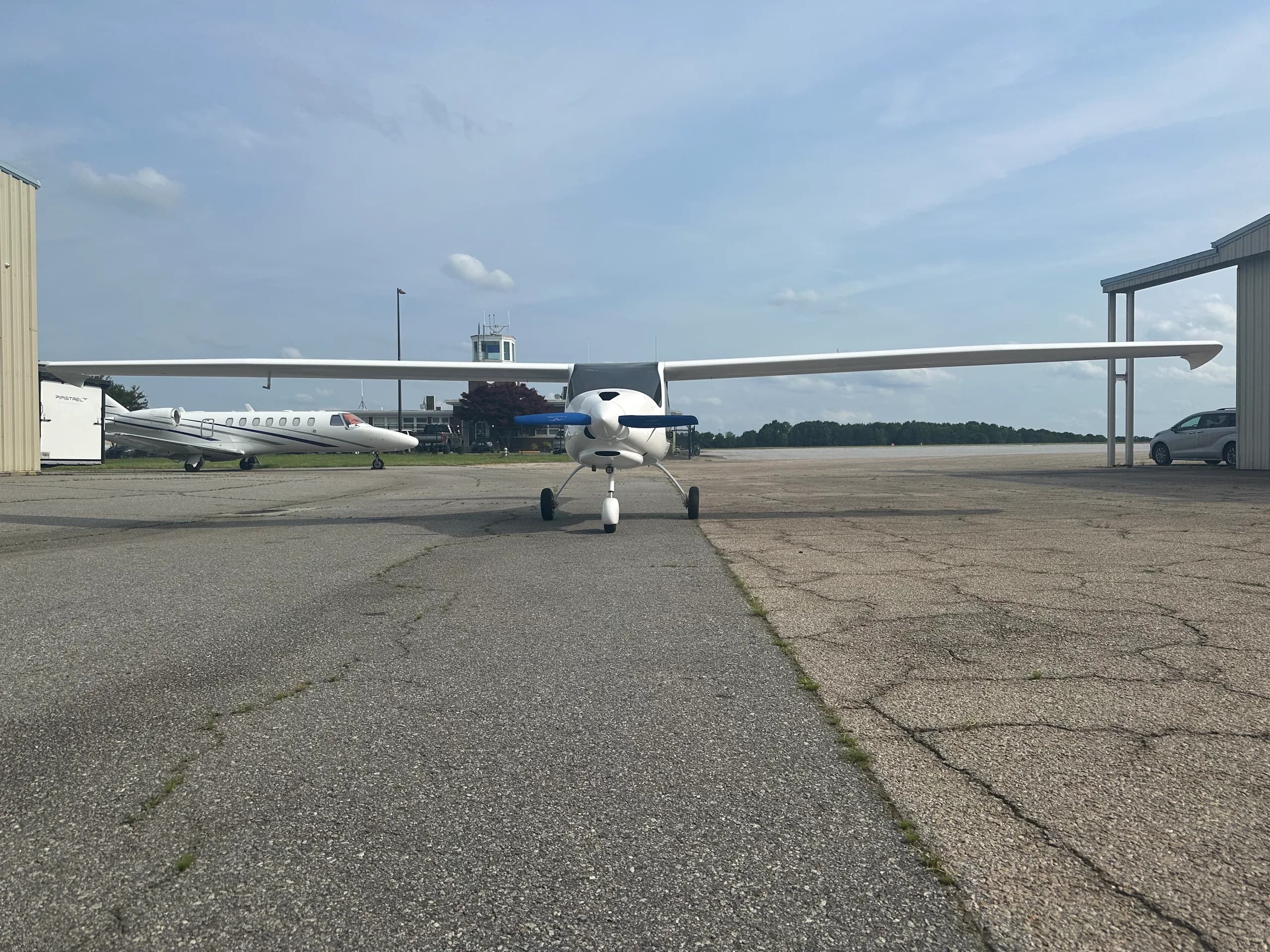 A white high-wing propeller aircraft with blue propeller tips is parked on an airport tarmac, with a jet and control tower in the background.