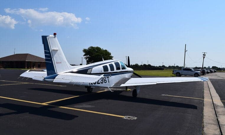 A white and blue Beechcraft Baron aircraft, N3236T, is parked on an asphalt tarmac with a building and cars in the distance.