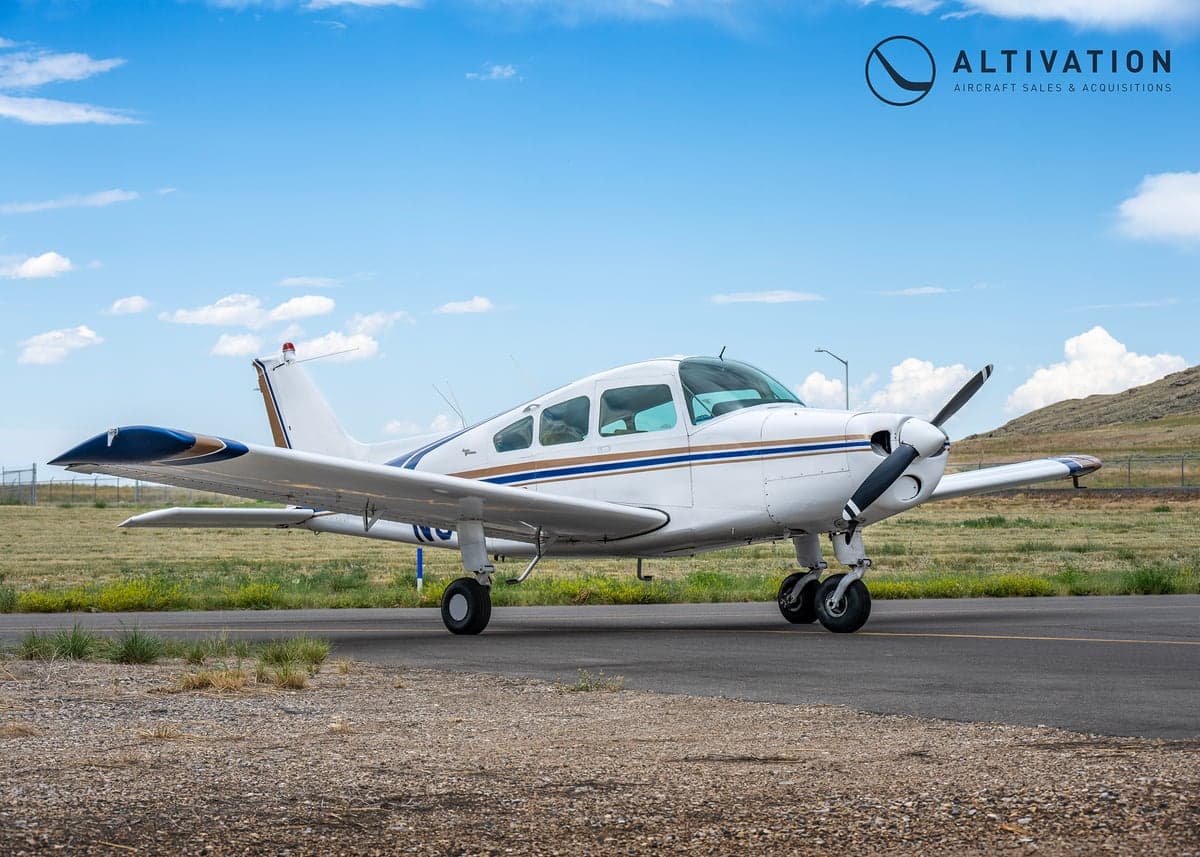 A white and blue Piper aircraft is parked on the tarmac on a sunny day.
