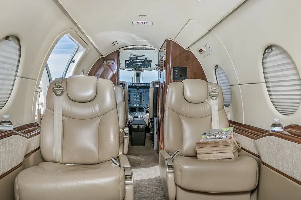 The interior cabin of a business jet with beige leather seats, wood trim, and a view towards the cockpit.