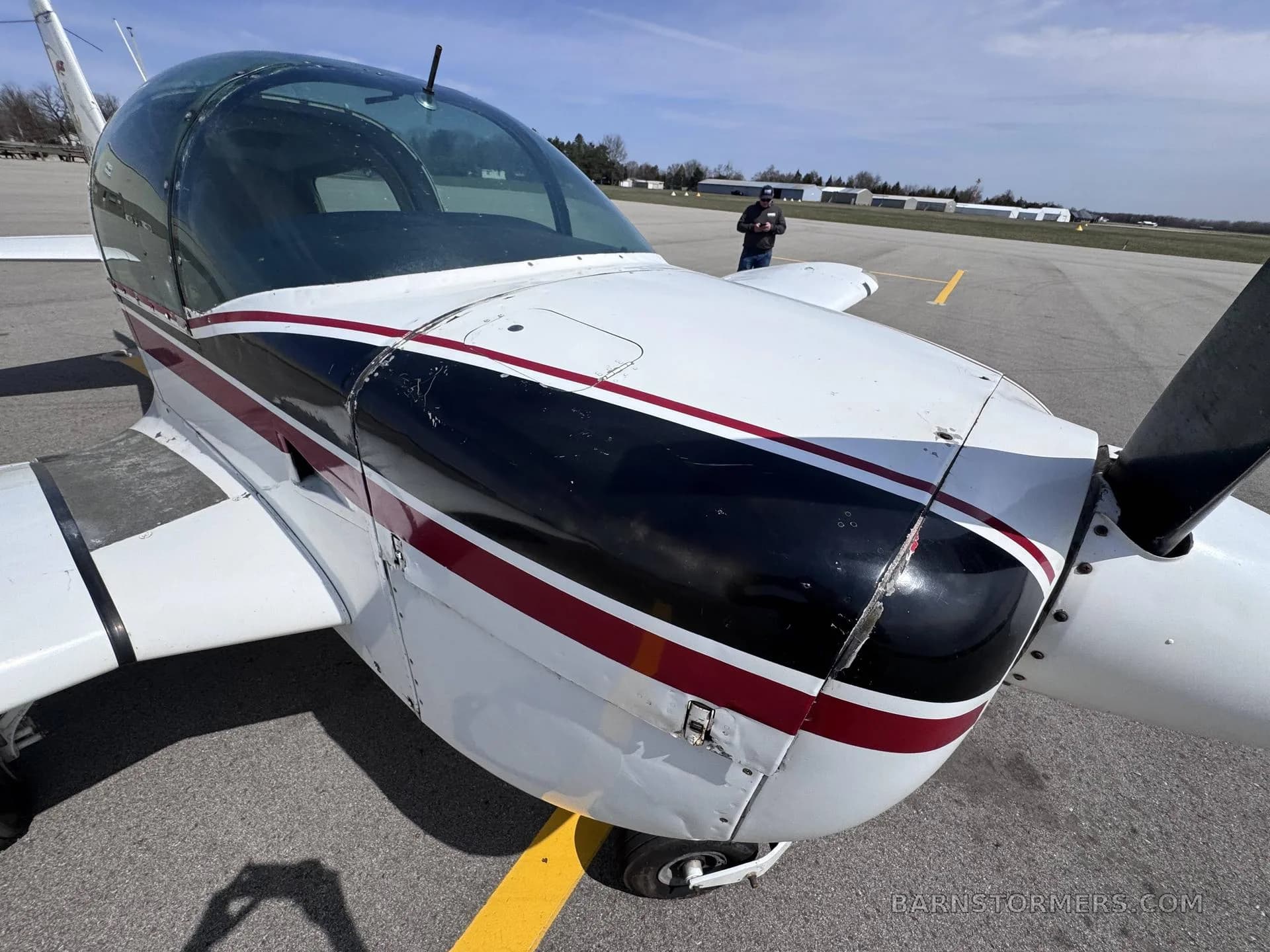 A close-up view of the front fuselage and propeller of a white and maroon Grumman American AA-5 Traveler aircraft.