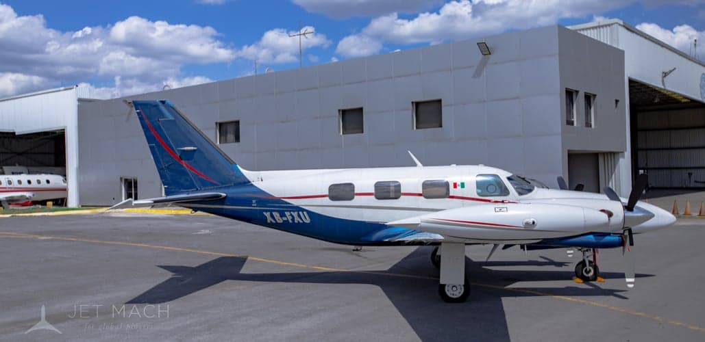 A white and blue twin-engine propeller aircraft, registration XB-FXU, is parked on a tarmac in front of a hangar.