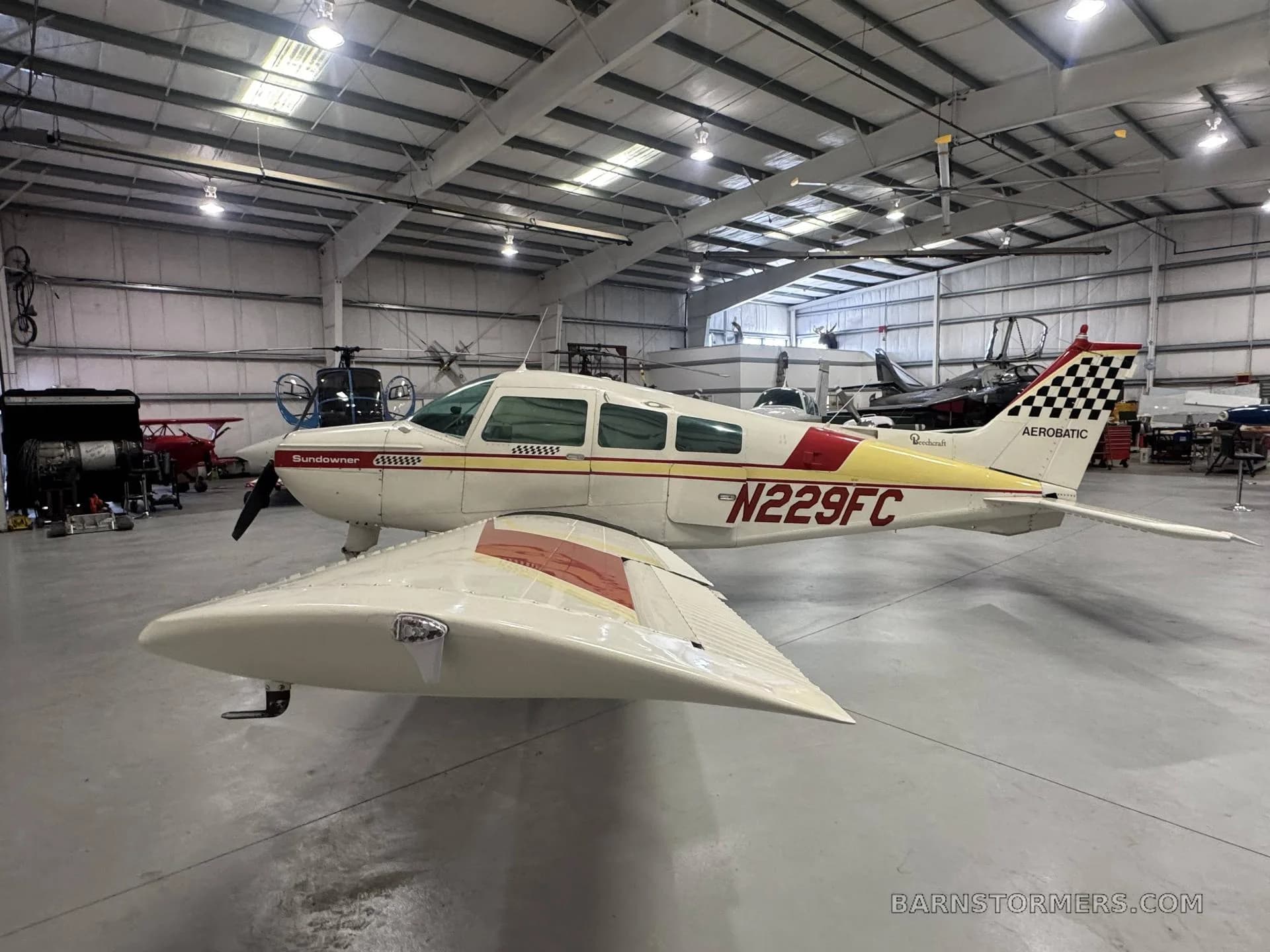 A white and red Beechcraft Sundowner N229FC with a checkered tail is parked in a hangar, viewed from the front-side.