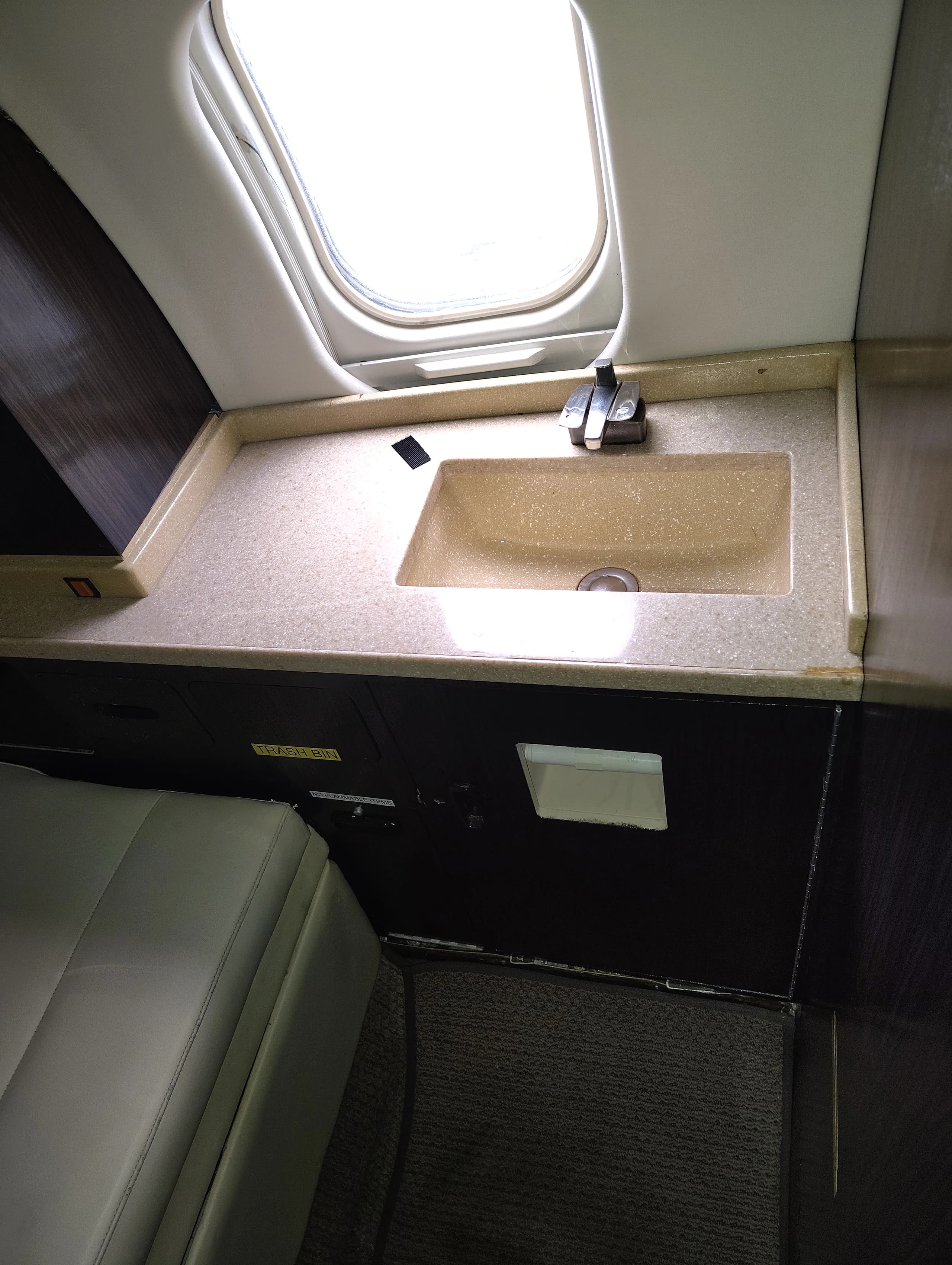 An aircraft lavatory or galley area features a beige countertop with a sink and faucet under a window.